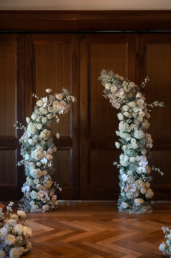 Wedding floral arch with white roses and orchids on floral pillars, set against a wood-paneled wall and parquet floor indoors