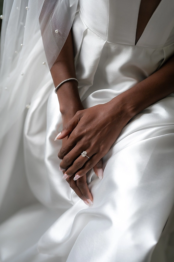 Engagement ring with an oval diamond engagement ring on bride’s hand, paired with a thin bracelet against soft satin gown fabric