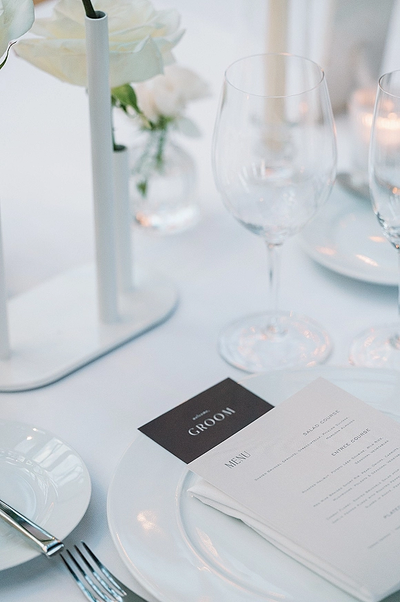 Wedding place setting with wedding menu card on a white napkin, stacked plates, silver flatware, wine glass, candle and white roses under soft lighting