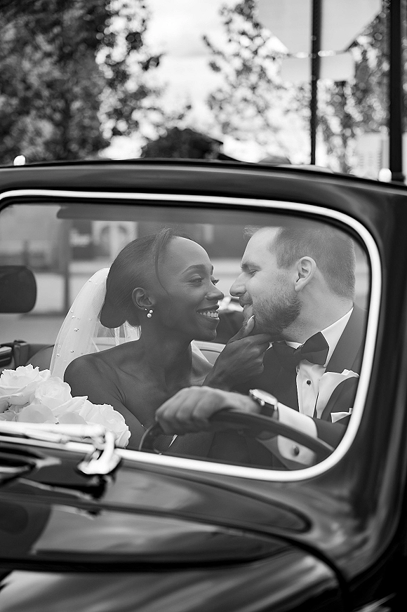 Wedding couple portrait of bride and groom in car, bride touching his face beneath her veil in a convertible, trees and street behind