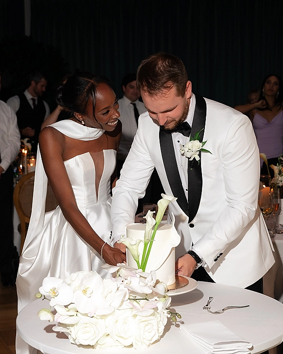 Wedding cake cutting as bride and groom slice a white cake topped with calla lilies and orchids at a candlelit reception table