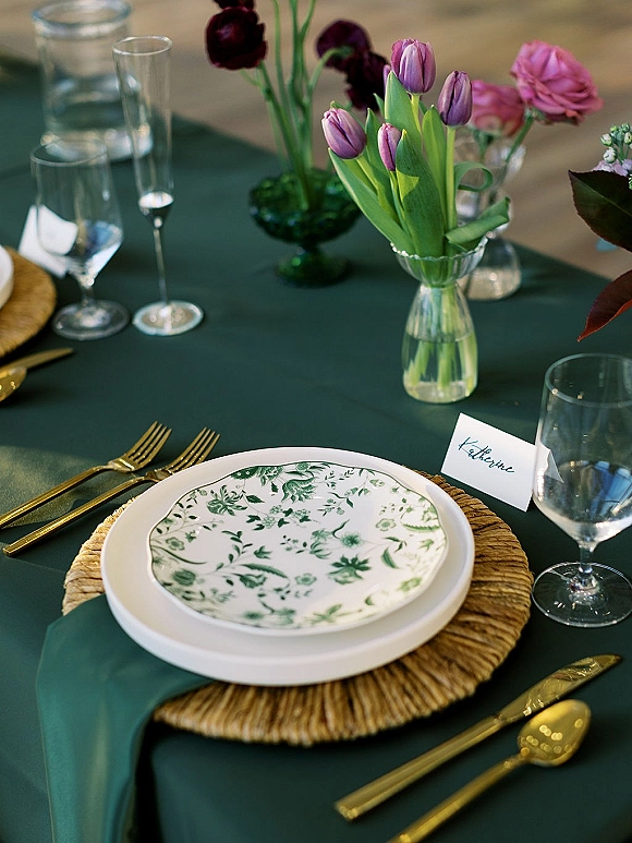 Reception tablescape with green wedding tablecloth, rattan charger, floral plate, gold flatware, and tulip centerpiece with pink flowers