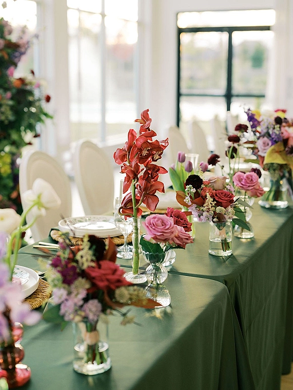 Reception tablescape with wedding table decor on a green tablecloth, orchids and roses in bud vases, rattan chargers and glassware by large windows