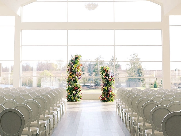 Ceremony setup with indoor wedding ceremony details, floral pillars and candle-lined aisle framed by white chairs and tall windows