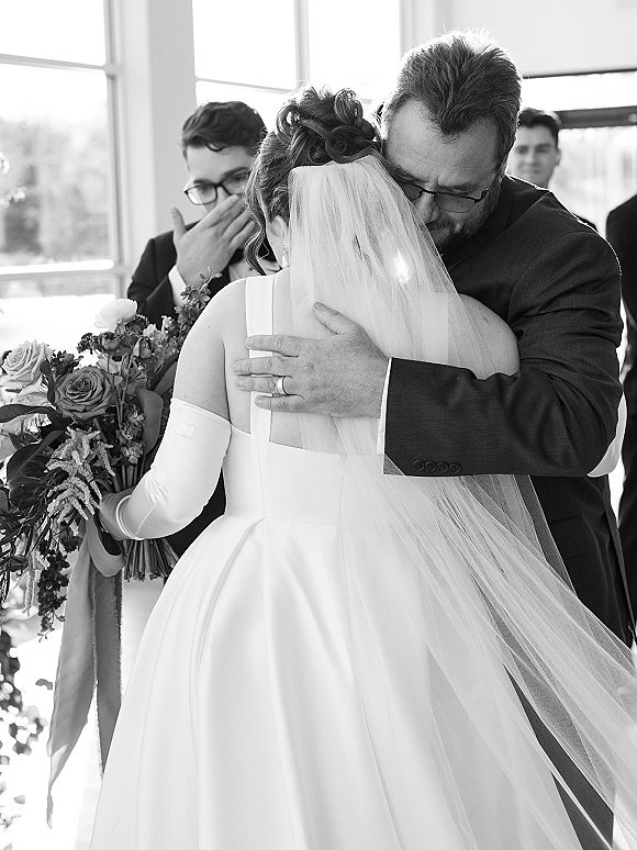 Father daughter wedding hug as the bride in veil and long gloves clutches a bouquet, embracing her dad in a suit by indoor windows with guests