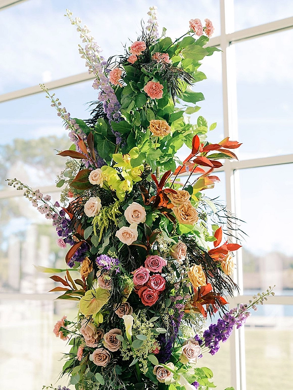 Wedding floral installation with a ceremony floral pillar of roses, orchids and calla lilies, glowing by large windows in natural light
