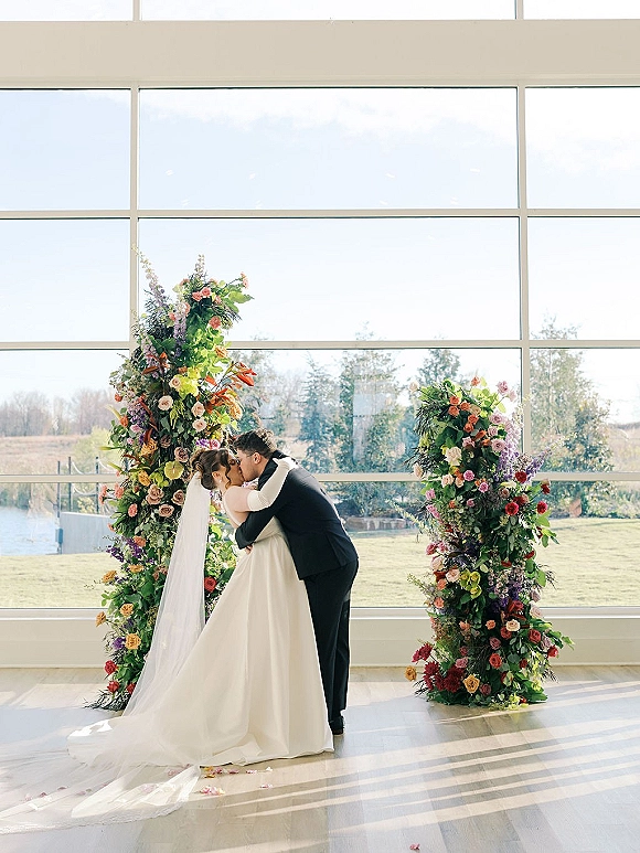 Wedding kiss portrait of bride and groom kissing under a colorful floral arch, veil flowing by floor-to-ceiling windows in daylight