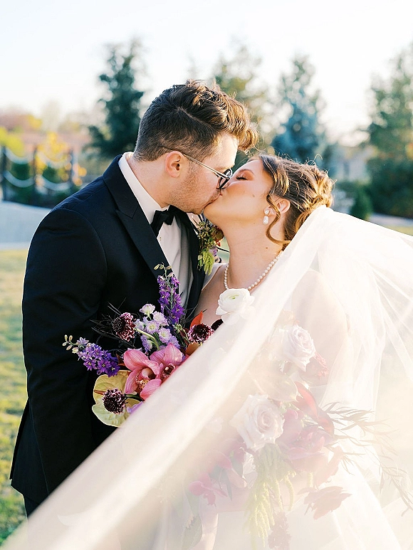 Wedding kiss as bride and groom kiss under a veil, her bouquet and pearls visible as he wears glasses and tuxedo on a sunny lawn