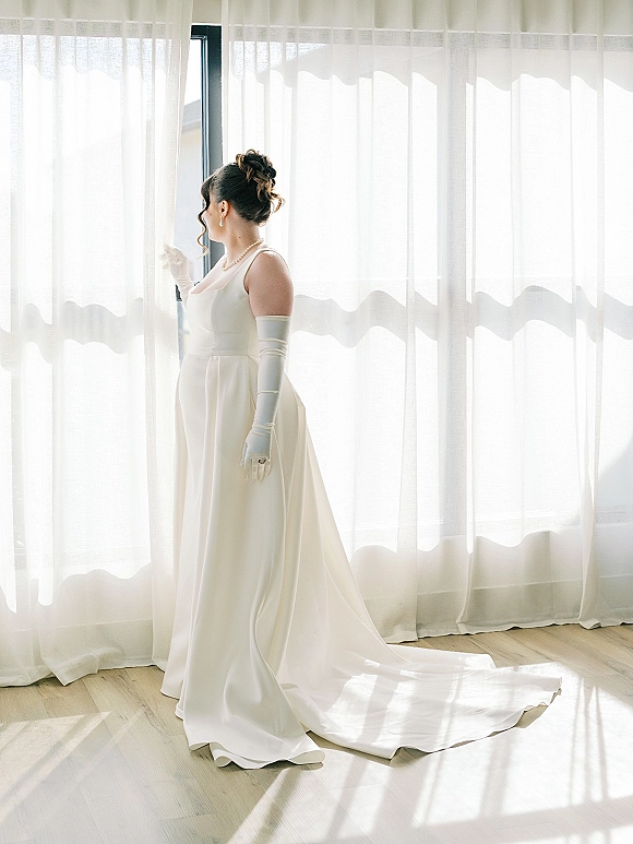 Bridal portrait of a bride by window in a simple satin wedding dress with opera gloves and pearl necklace, framed by sheer curtains