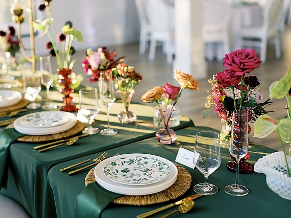 Reception tablescape with green wedding tablecloth, patterned plates, woven placemats, gold flatware, pink florals, and taper candles in a bright room