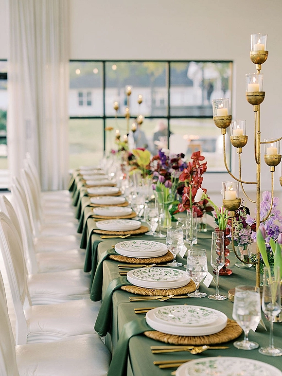 Reception tablescape on a long banquet table with green table linen, patterned plates, rattan chargers, gold flatware, and tall candelabra by grid windows.