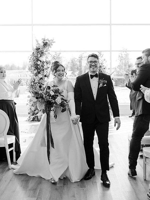 Wedding recessional as bride and groom walk the aisle holding hands, bride in veil with bouquet ribbons, guests in bright windowed venue