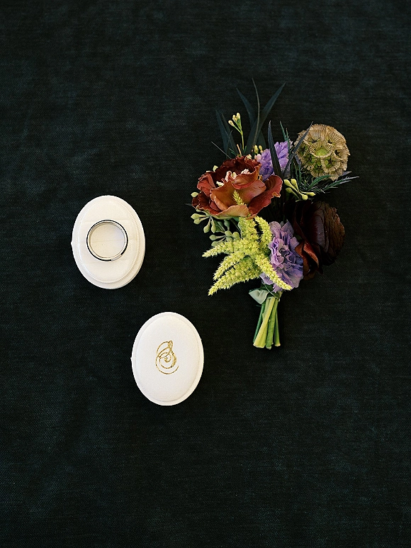 Wedding ring boxes in white beside an engagement ring, wedding band, and dark red and purple bouquet on a black fabric backdrop