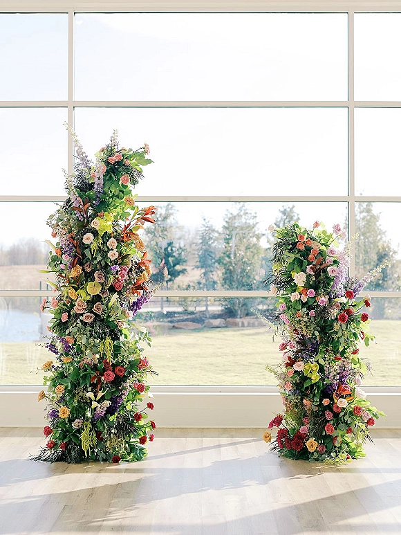 Wedding ceremony arch with an asymmetrical floral arch of colorful roses and greenery beside floor-to-ceiling windows overlooking lawn and water view