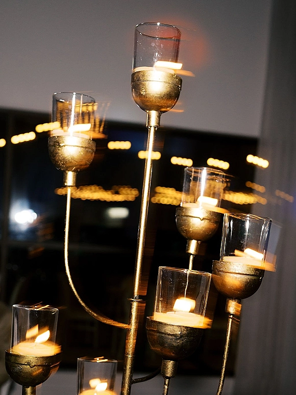 Wedding candle centerpiece with a gold metal candelabra and glass hurricane shades, glowing with votives in a dark room with string light bokeh