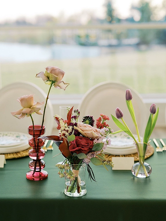 Reception tablescape with wedding table centerpiece of roses and tulips in a clear vase, red candlestick, and place cards by a window view