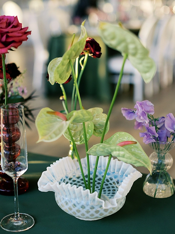 Wedding centerpiece featuring anthurium centerpiece stems with burgundy roses and sweet peas in a glass vase on dark green linen at an indoor reception table