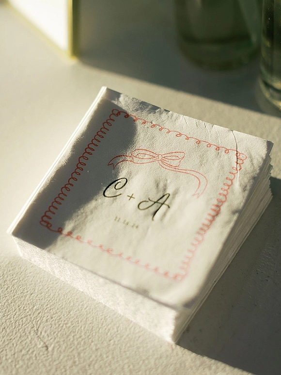 Wedding cocktail napkins with custom monogram initials, ribbon illustration, and red border on a sunlit tabletop beside drinking glasses