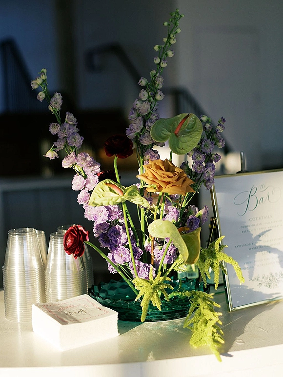 Cocktail bar decor with wedding bar sign, teal glass bowl of anthurium and ranunculus beside acrylic cups and cocktail napkins on counter