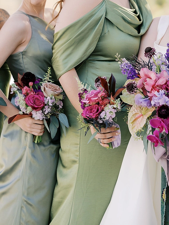 Bridesmaid bouquets held in front of sage green bridesmaid dresses, colorful roses and orchids with eucalyptus in soft outdoor light