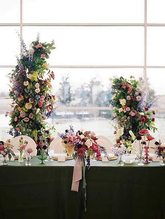 Reception tablescape with head table flowers on a green tablecloth, taper candles and bud vases, set by large windows with trees outside