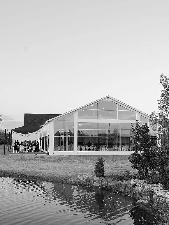 Wedding reception venue with a glass wall event space, string lights over outdoor dining tables beside a pond, lawn, and trees under open sky