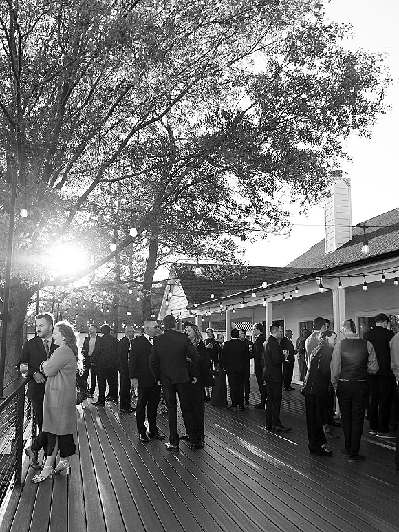 Cocktail hour guests mingling at a wedding cocktail hour on a wood deck, holding drinks under string lights at sunset by a house porch