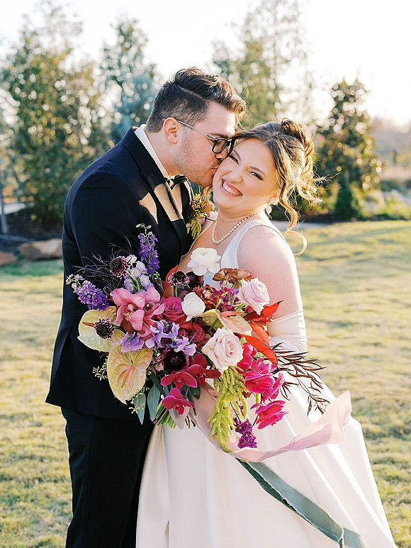 Wedding couple portrait of groom kissing bride’s cheek as she laughs, holding a colorful bouquet on a sunlit garden lawn with trees