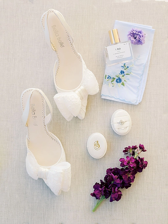 Bridal details flatlay with wedding shoes flatlay, white lace bow heels, perfume, embroidered handkerchief, rings, and a purple flower on linen