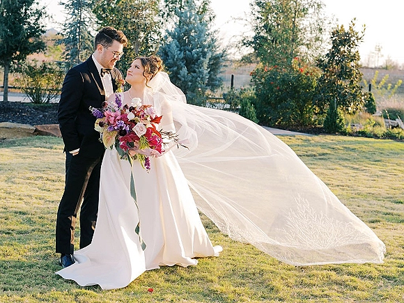 Couple portrait of bride and groom holding a colorful bouquet, her long veil flowing in sunlight on a grassy lawn with trees and hillside