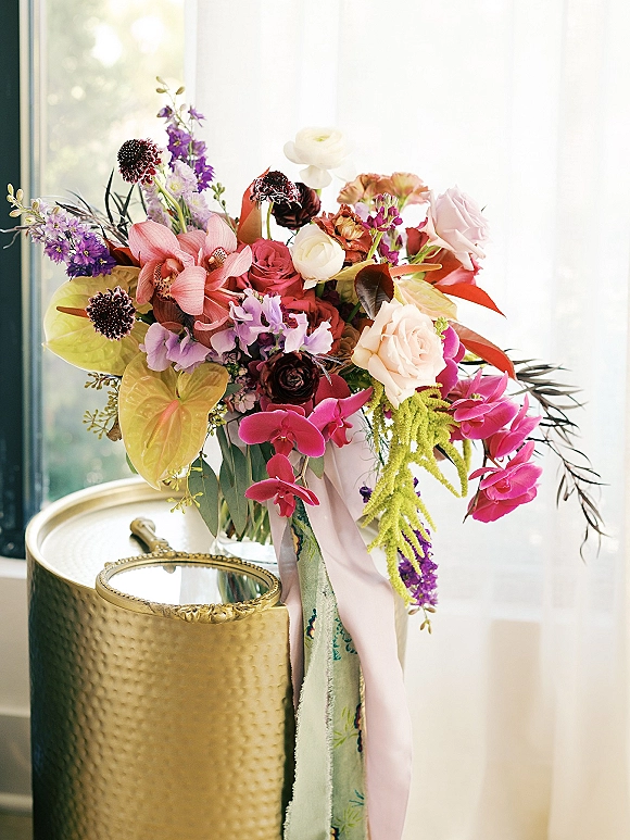 Wedding bouquet with colorful bridal bouquet blooms of orchids, roses, and anthurium in a glass vase by window light on a gold side table