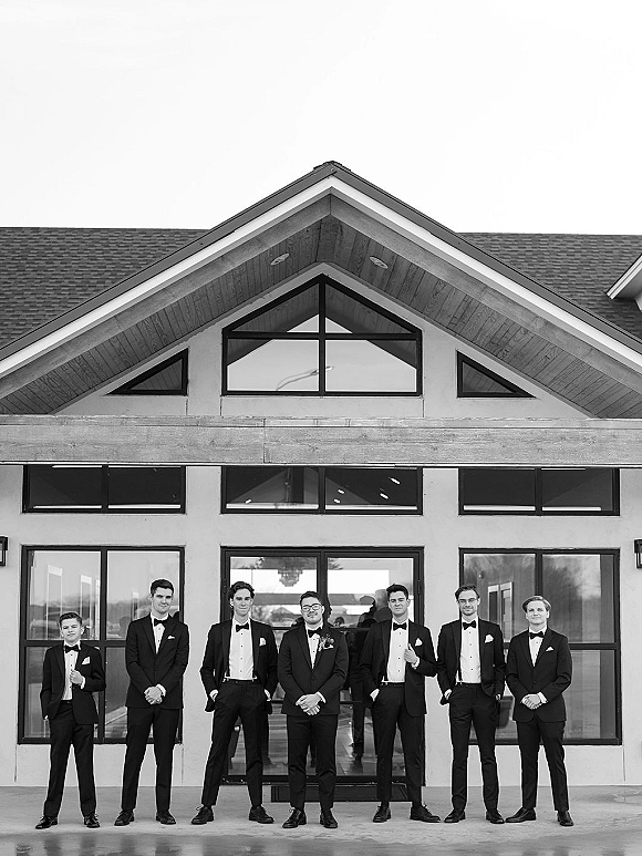 Groomsmen portrait of men in tuxedos with bow ties and boutonnieres lined up on a concrete patio outside a modern glass venue