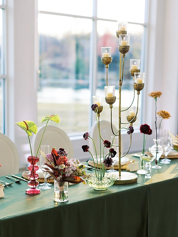 Reception tablescape with a green wedding tablecloth, tall candelabra centerpiece, pillar candles, and florals by a large window overlooking scenery