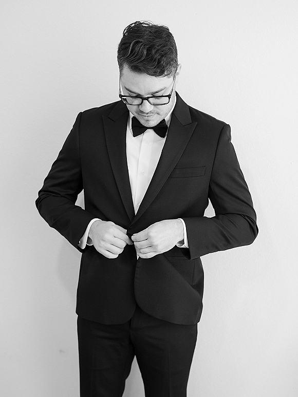 Groom portrait in a black tuxedo with bow tie and eyeglasses, standing against a plain wall for a minimalist, classic look