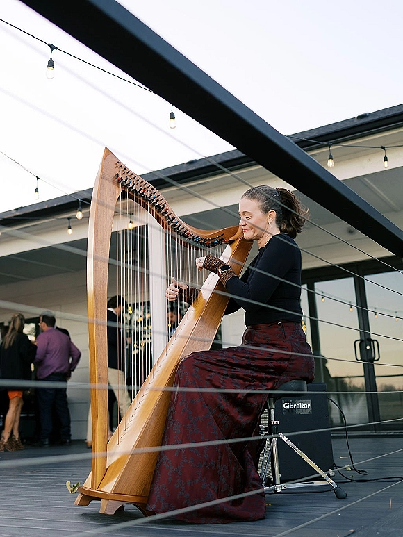 Wedding live music with a wedding harpist performing on a patio stage under string lights, with guests mingling by a modern venue