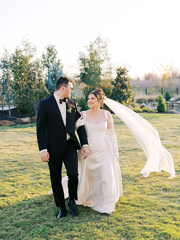 Couple portrait of bride and groom walking hand in hand on a lawn, her long veil blowing in wind as she looks at him