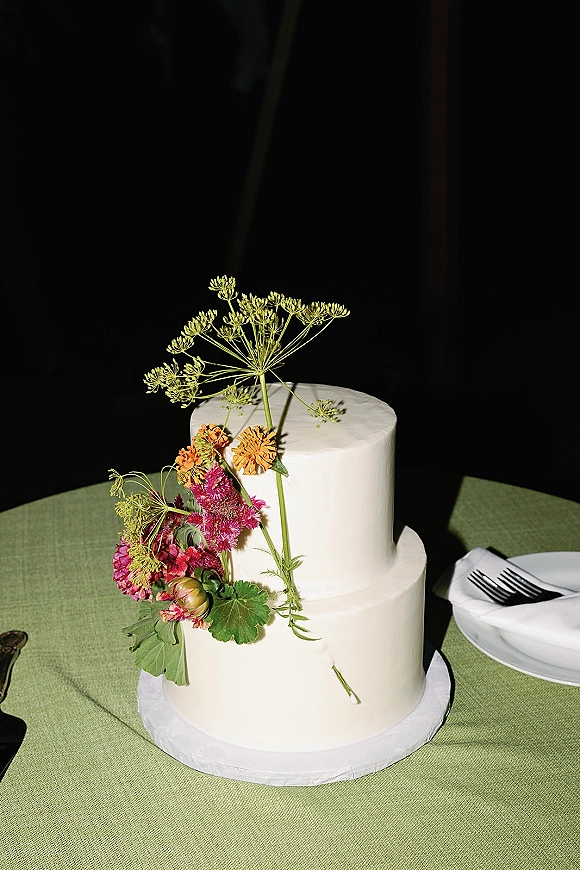 Wedding cake with a two tier wedding cake design, smooth white buttercream, and fresh wildflower topper on a green-clothed table against dark backdrop