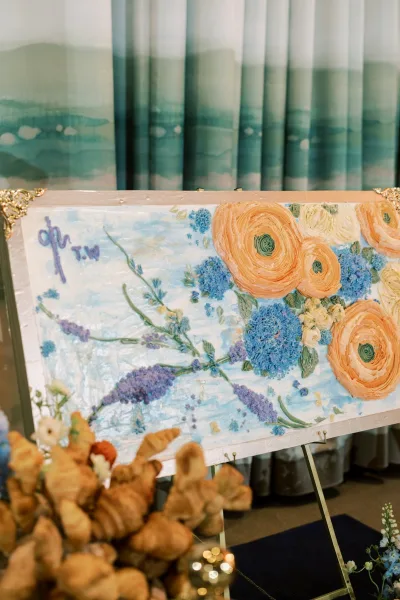 Blue and peach floral artwork in a gold frame stands on an easel beside croissants and draped curtains