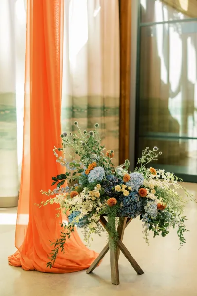 Orange and blue hydrangeas, orchids and mums make up the ceremony flowers against sheer draped fabric