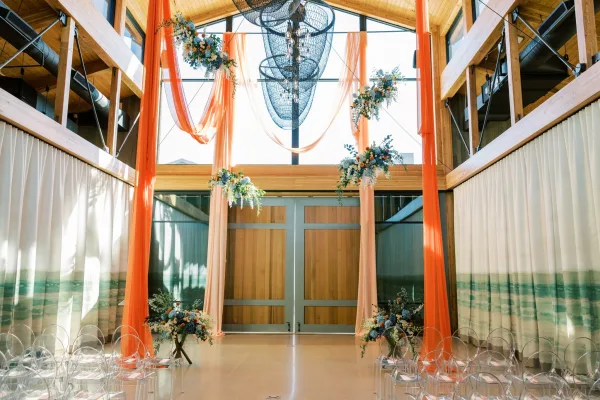Orange draping and suspended blue-and-peach flowers frame the ceremony setup, with acrylic ghost chairs beneath a chandelier and wood beams