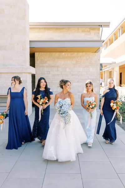 Bride and bridesmaids walk through a stone courtyard in navy and light-blue dresses, carrying orange bouquets