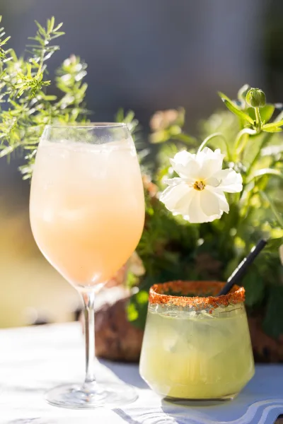 Salt-rimmed wedding cocktails with ice cubes and a flower garnish sit on a white tablecloth amid garden greenery