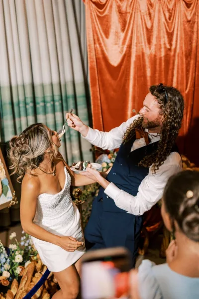 Newlyweds feed each other a slice of chocolate cake, with the bride wearing a beaded mini dress