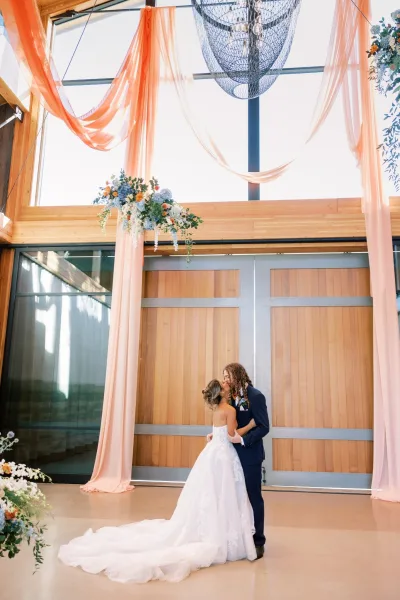 Bride and groom kiss beneath blue and white florals, framed by peach drapery and a statement chandelier