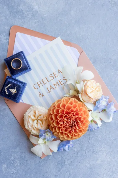 Blue-and-peach striped wedding invitation suite with gold foil lettering beside a blue velvet ring box on a textured surface
