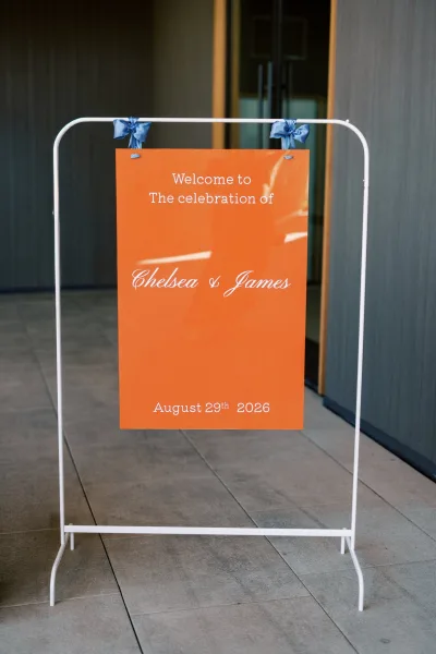 Orange wedding welcome sign with script and serif lettering, tied ribbons and a white metal stand before glass doors
