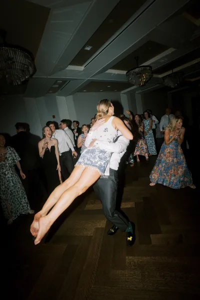 Wedding reception dancing as a groom lifts the bride on a crowded ballroom dance floor, guests cheering under chandeliers with champagne flute accents