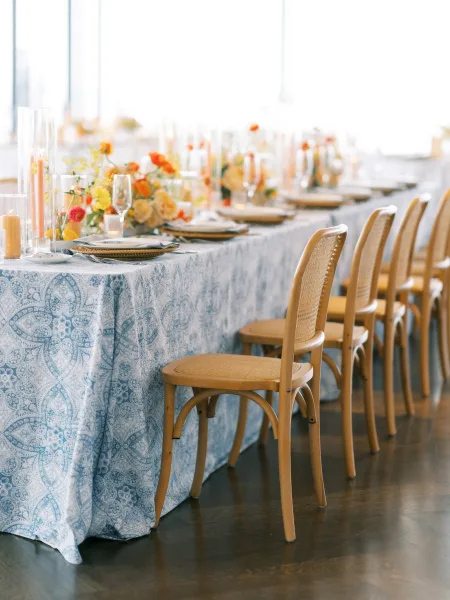 Reception tablescape on a long banquet table with a blue and white tablecloth, orange-yellow florals, taper candles, and rattan chairs by large windows