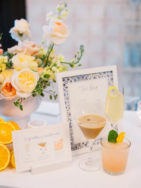 Signature cocktail bar with signature cocktail menu cards, mixed drinks, and citrus garnish on a white tabletop against a neutral wall