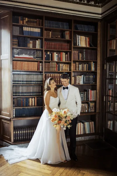 Couple portrait of bride holding a bouquet and groom in a white dinner jacket, laughing together before library bookshelves in window light